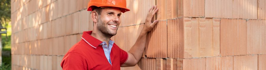 Muž stojící před rohem cihlové stěny z cihel Porotherm T Profi na hlavě s oranžovou helmou rukou hladící cihly